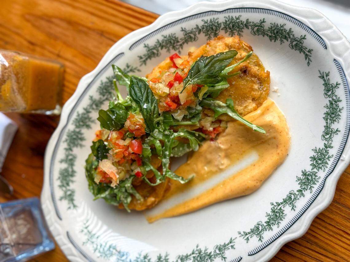 A close-up, high-angle shot of a gourmet appetizer of fried green tomatoes, served on an ornate, oval-shaped white plate with a green floral border. The golden-brown fried tomatoes are topped with a fresh, colorful chopped relish and arugula, and are served with a smear of a creamy, light-orange sauce on the plate. The dish is presented on a slatted wooden table, with a bottle of hot sauce blurred in the top-left corner.