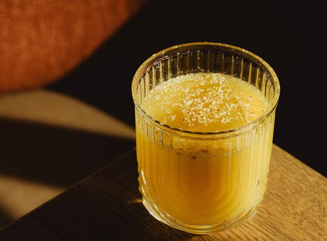 A close-up of the Picante Infante cocktail in a textured, ribbed glass. The drink is a vibrant yellow-orange color, served over a large, clear ice cube and topped with a fine dusting of spices. The glass sits on a wooden surface, illuminated by a warm, direct light that creates a dramatic shadow play in the background.