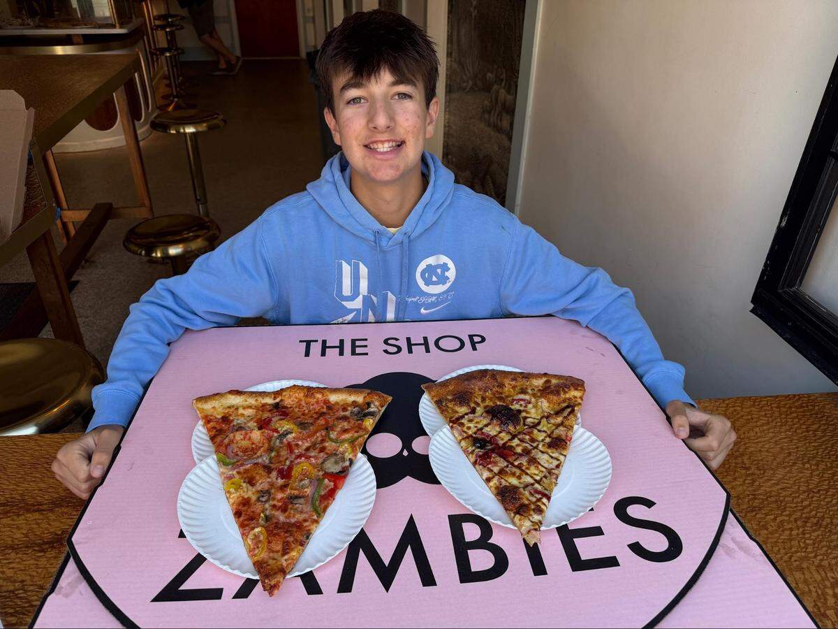 An eye-level, medium shot of a smiling teenager sitting behind a large pink pizza box that functions as a tabletop. He is wearing a light blue UNC Chapel Hill hooded sweatshirt. Resting on a box in front are two white paper plates, each holding an oversized slice of New York-style pizza—one loaded with various vegetable toppings and the other drizzled with a crisscross pattern of dark barbecue sauce. The pink surface features a black skull logo and reads “THE SHOP ZÄMBIES.”