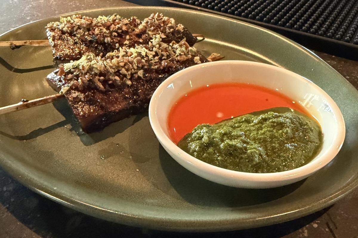Two grilled skewers are served on an olive-green ceramic plate alongside a vibrant dipping duo, with one half a bright, fiery orange chili oil or harissa, and the other half is a thick, textured green herb salsa or chimichurri.