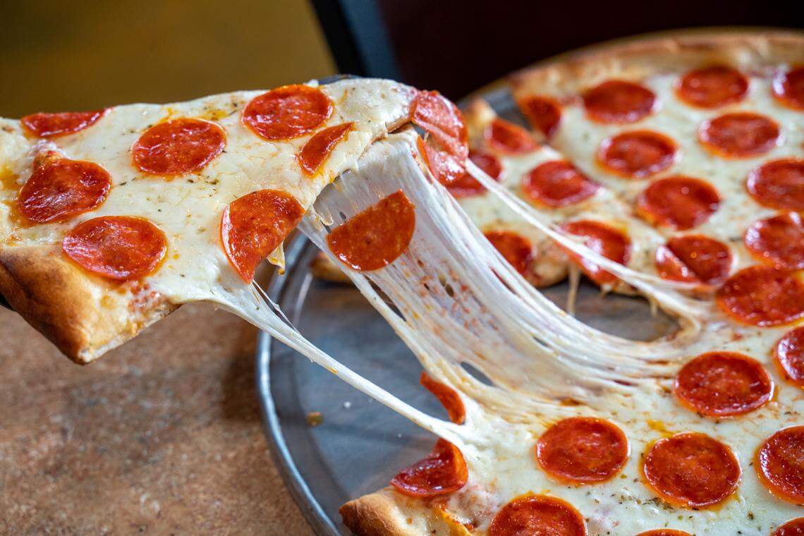 A slice of pepperoni pizza is lifted from the pie, creating a long, dramatic pull of stretching, melted mozzarella cheese. The rest of the pizza sits on a metal pan in the background.