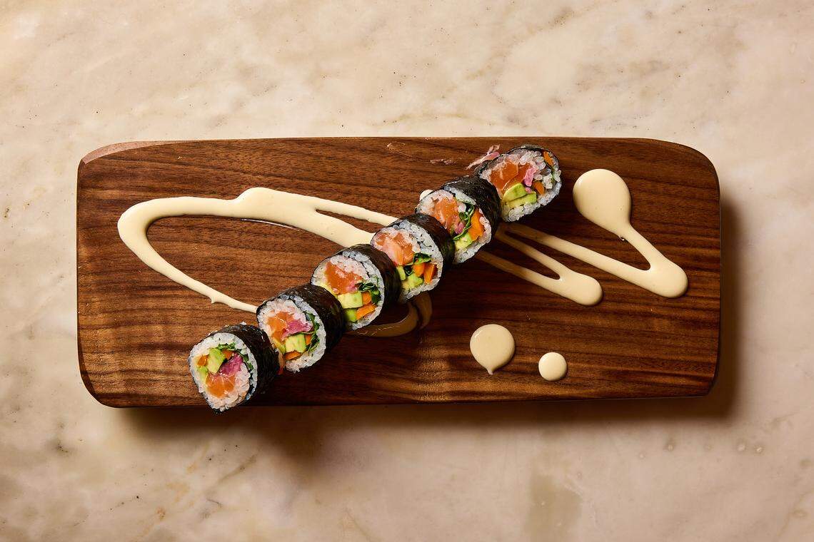 An overhead shot of a sushi roll cut into six pieces, arranged in a diagonal line on a long, dark wood serving board. The roll contains salmon, avocado, and vegetable julienne. The board is decorated with artistic swooshes and dots of a creamy, pale yellow sauce. The entire setting rests on a light-colored marble surface.