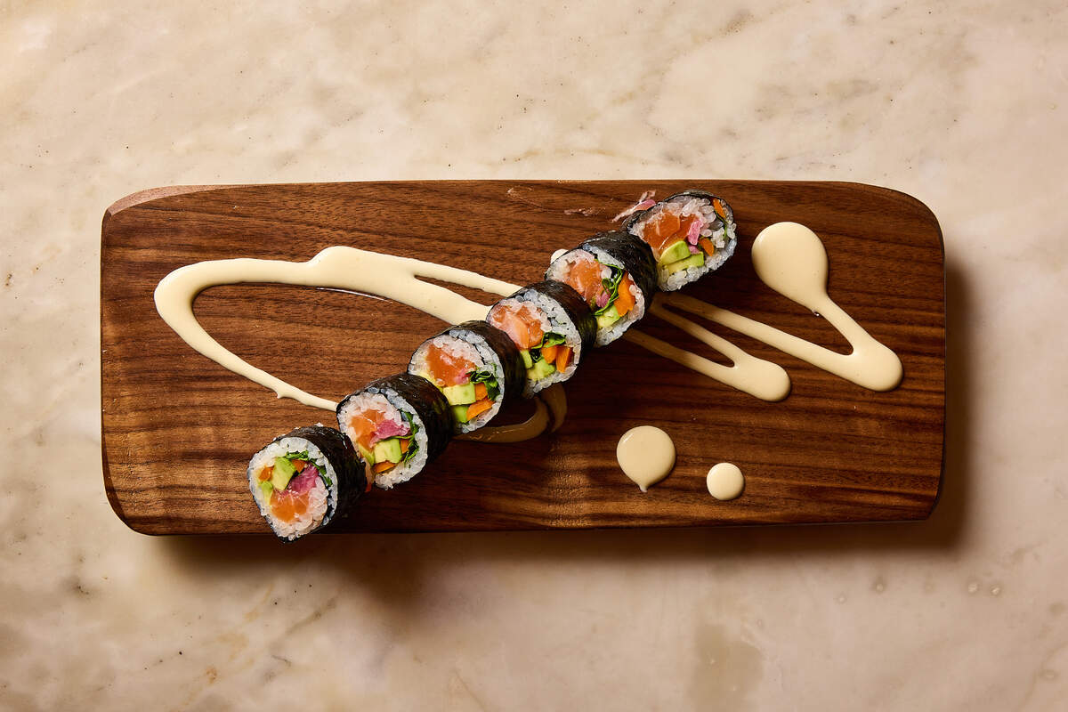 An overhead shot of a sushi roll cut into six pieces, arranged in a diagonal line on a long, dark wood serving board. The roll contains salmon, avocado, and vegetable julienne. The board is decorated with artistic swooshes and dots of a creamy, pale yellow sauce. The entire setting rests on a light-colored marble surface.