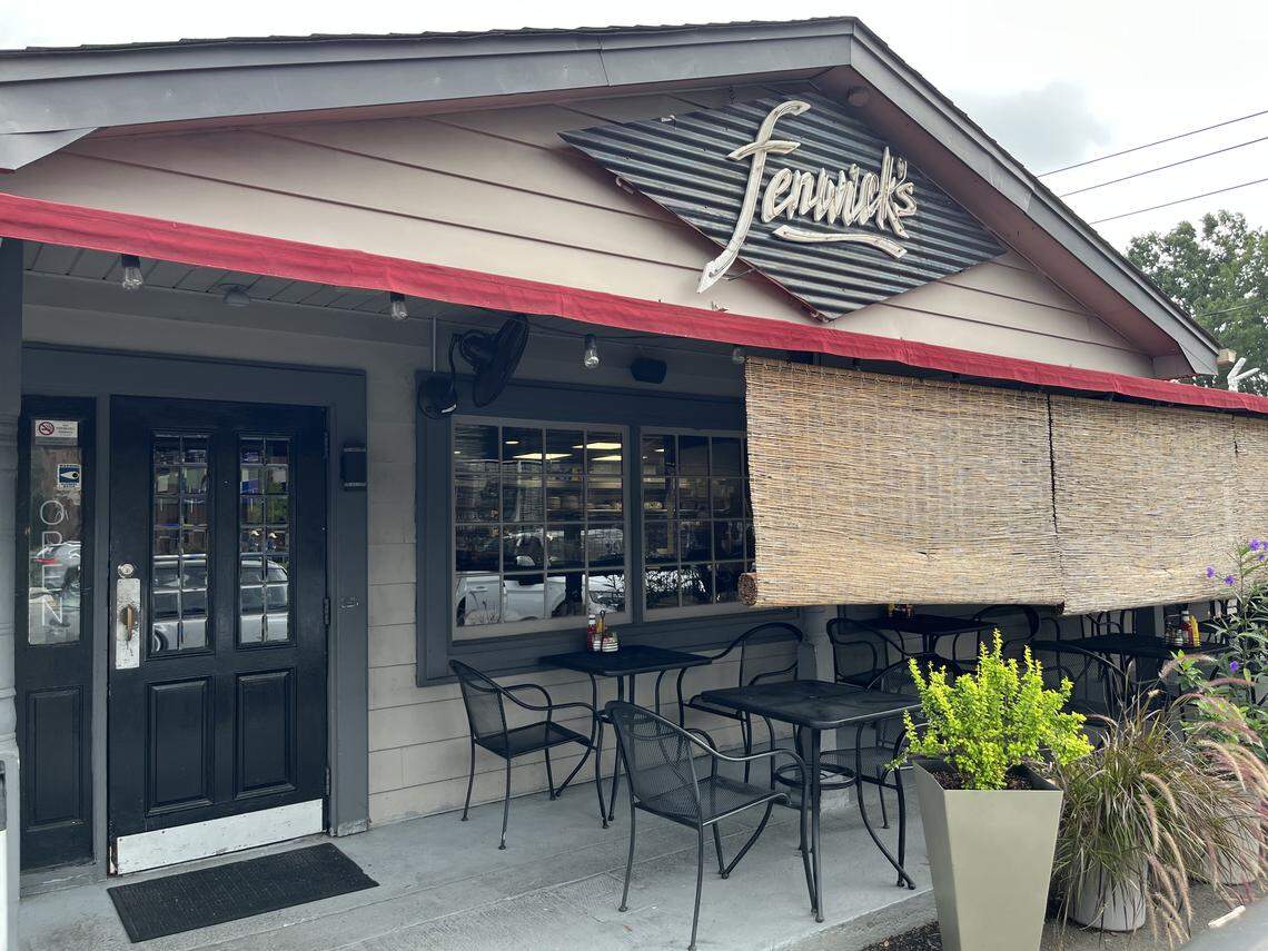 A street-level view of the entrance to Fenwick’s restaurant. The single-story building has tan siding and a dark grey front door. A red awning stretches over a small concrete patio with black metal tables and chairs. Above the awning, the name “Fenwick’s” is written in a stylized font on a tilted, diamond-shaped sign made of corrugated metal.
