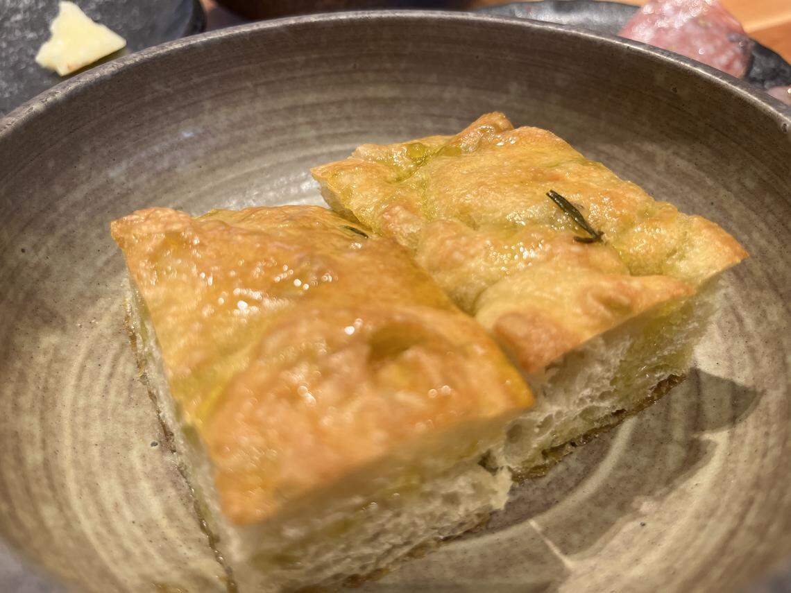A close-up shot of two rectangular pieces of golden-brown focaccia bread, glistening with olive oil and topped with a sprig of rosemary. The bread, which has a soft-looking interior, is served in a round, rustic gray bowl. In the blurred background, other dishes and a piece of white cheese are visible.