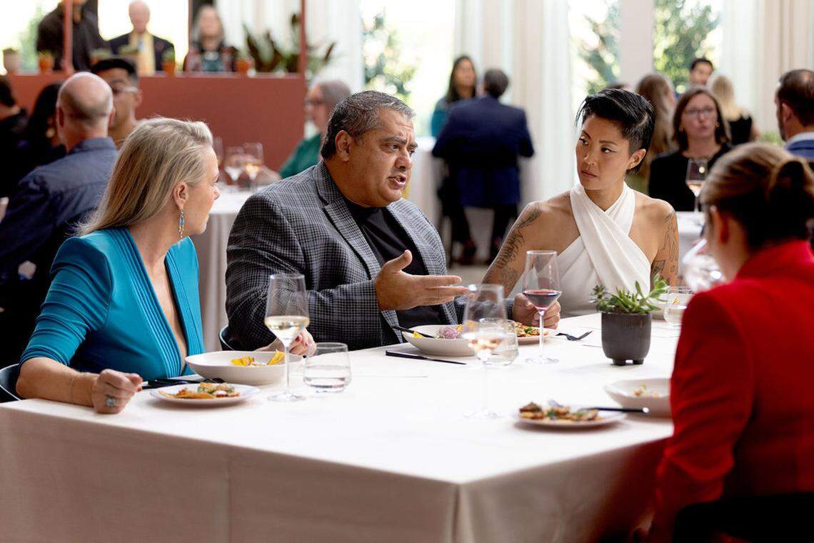 A group of well-dressed guests engaged in conversation at a white-clothed dinner table. A diner in a grey patterned blazer gestures while speaking to two others —one in a bright turquoise dress and another in a white halter top with visible arm tattoos. Wine glasses and plated appetizers are set before them in a bright, airy venue.