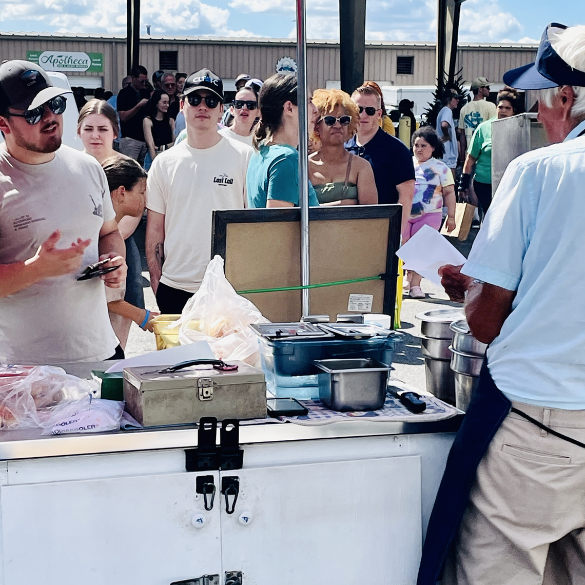 A vendor wearing a white visor and a light blue shirt stands behind a food cart, talking to a customer and holding a piece of paper. The customer, in a white t-shirt and sunglasses, is also speaking and holding a phone. A line of people is waiting behind them, including a customer with red hair and sunglasses, and another person in a white t-shirt with a “Lost Sail” logo. The food cart has a variety of items on it, including containers, a cash box, and bags. The scene appears to be an outdoor market or event under a covered awning on a sunny day.