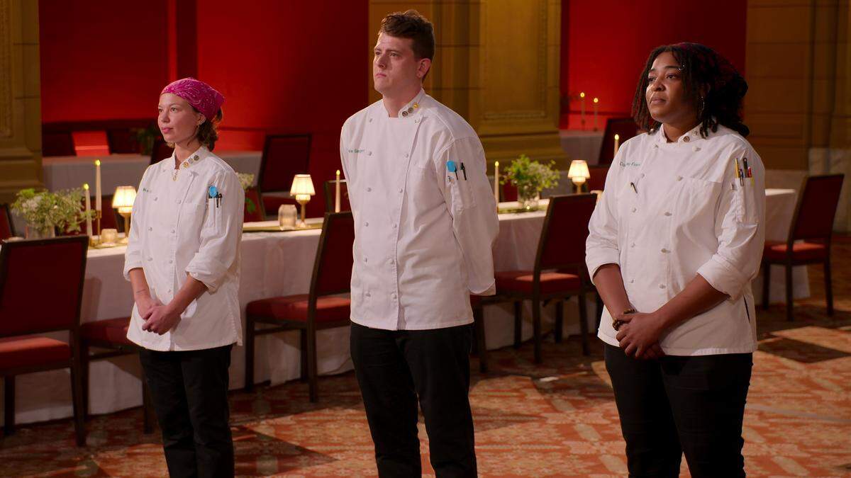 Three chefs stand in a line facing forward with serious, expectant expressions. They are dressed in matching white chef’s coats, appearing as contestants in a cooking competition. The setting is an elegant dining hall, with a long, formally set table with lit candles and lamps visible behind them.