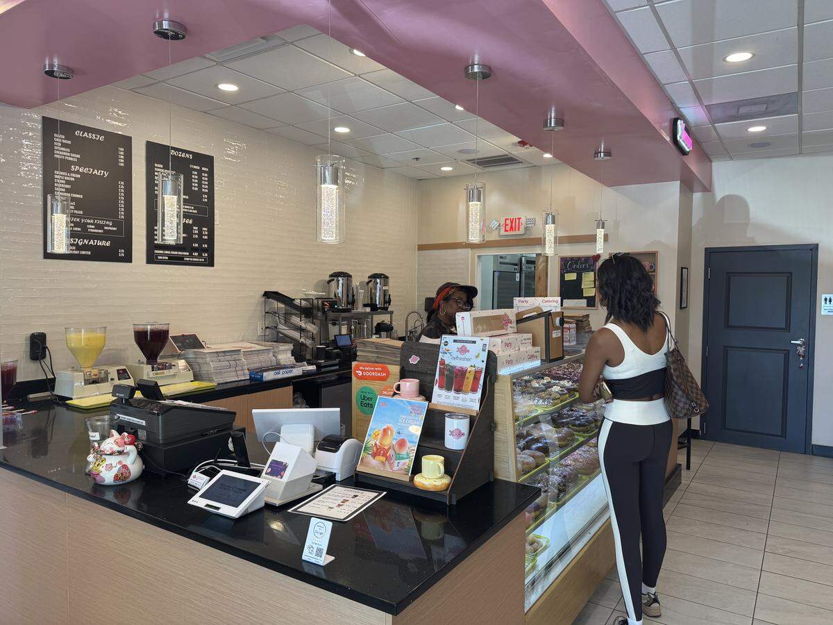 A customer in black leggings and a white and black top is standing in line at a doughnut shop, looking at a display case filled with assorted doughnuts. The counter in front of the display case is black with a menu hanging on the wall behind it. A worker wearing a black shirt and red hat stands behind the counter.
