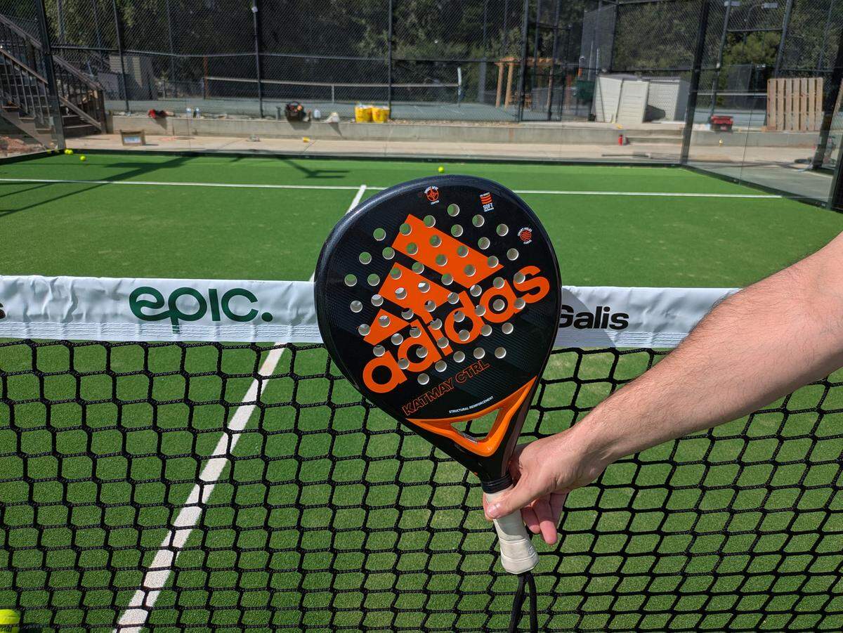 A person holds a black padel racket with a bright orange Adidas logo in the foreground. The racket is positioned in front of a black net on a green court with white lines. The background shows another court and a chain-link fence.