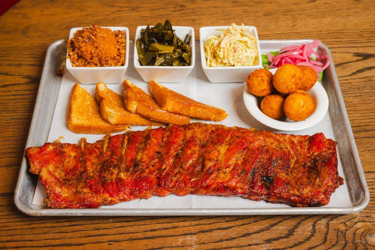A metal tray holds a long, glazed rack of ribs alongside four slices of triangular Texas toast and a small bowl of golden-brown hushpuppies with pickled red onions. Three small white square bowls contain southern sides: sweet potato mash with a crumble topping, collard greens, and creamy coleslaw.