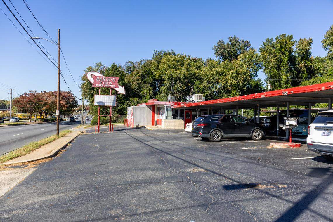 A wide, eye-level shot of the vintage “Bar-B-Q King” drive-in restaurant. The view from the parking lot shows the iconic red and white “CURB SERVICE” sign with its large arrow, the small red and white building, and the long red canopy covering parked cars. The scene is set on a sunny day, with a multi-lane road to the left and a backdrop of dense green trees.