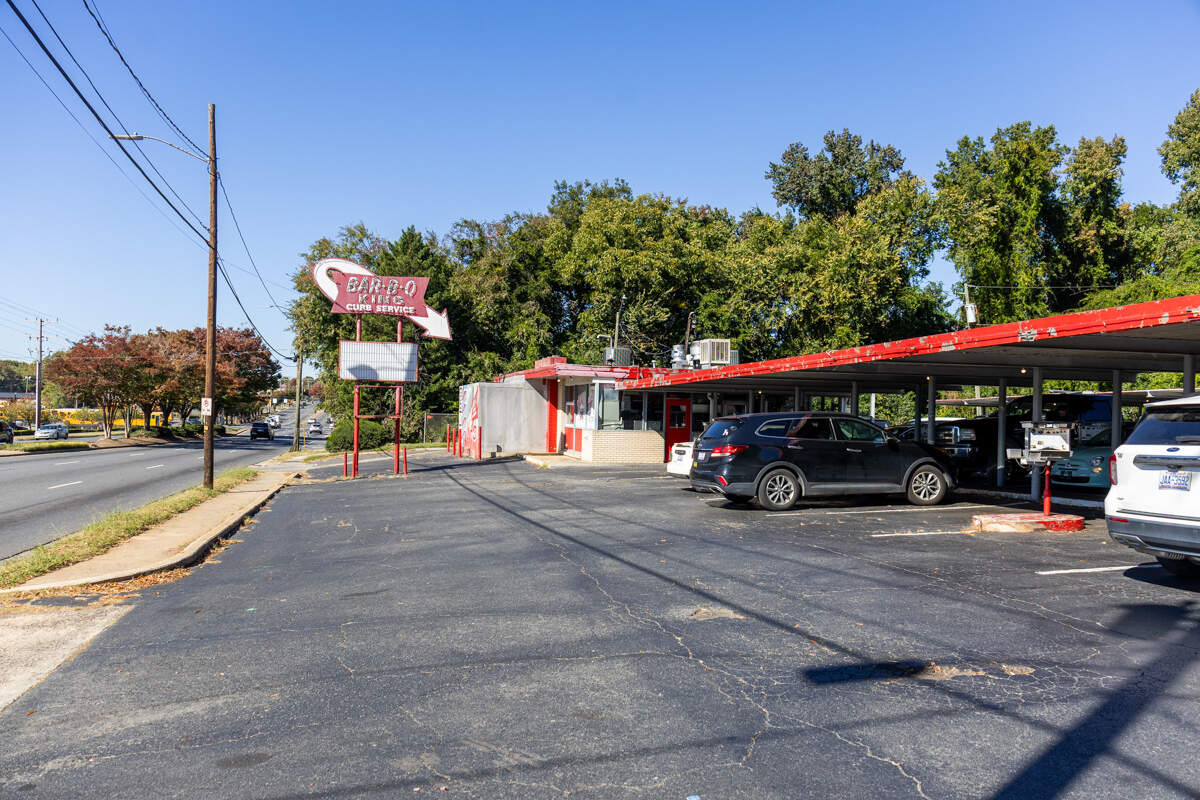 A wide, eye-level shot of the vintage “Bar-B-Q King” drive-in restaurant. The view from the parking lot shows the iconic red and white “CURB SERVICE” sign with its large arrow, the small red and white building, and the long red canopy covering parked cars. The scene is set on a sunny day, with a multi-lane road to the left and a backdrop of dense green trees.