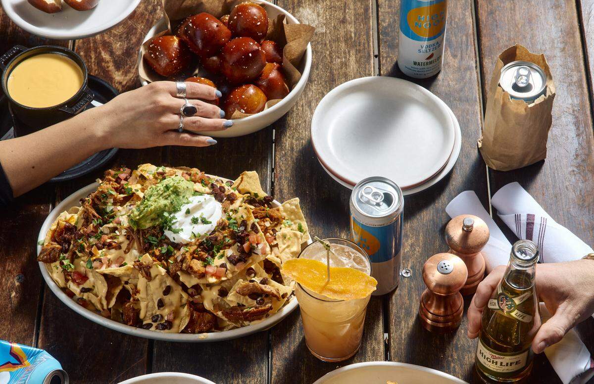 A top-down view of a wooden table filled with pub favorites, including a large plate of loaded nachos, pretzel bites, and various drinks. A person’s hand reaches for the pretzel bites, capturing a social and casual dining atmosphere.