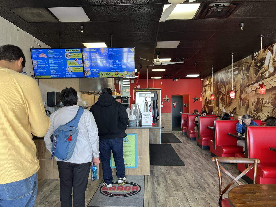 An eye-level, wide shot of the interior of a Sabor Latin Street Grill. On the left, a line of customers waits to order at a counter beneath bright blue digital menu boards. To the right, diners are seated in red vinyl booths along a wall featuring a large, sepia-toned mural, while the back wall is painted a vibrant red.