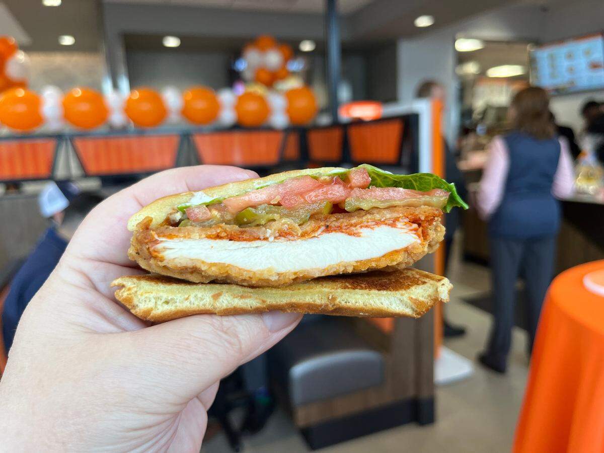 A person holds up half of a spicy chicken sandwich to the camera. The cross-section shows a thick, white-meat fried chicken fillet, green jalapeño slices, and red tomato between two pieces of thick, buttery toast. The background is the interior of a bustling restaurant decorated with orange balloons.