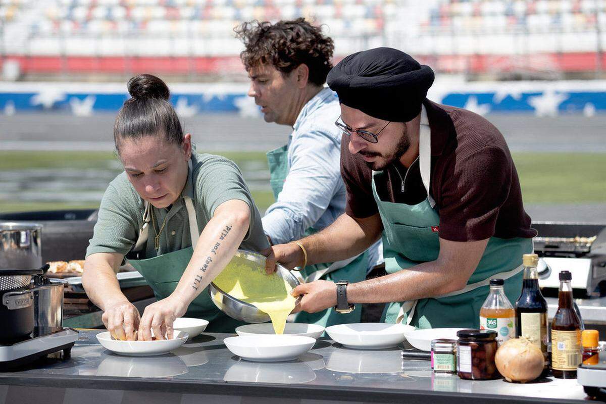 Two chefs in green aprons are focused on food preparation. One chef is pouring a yellow liquid from a metal bowl into white plates while another chef with a “I’ll keep you wild” tattoo works nearby.