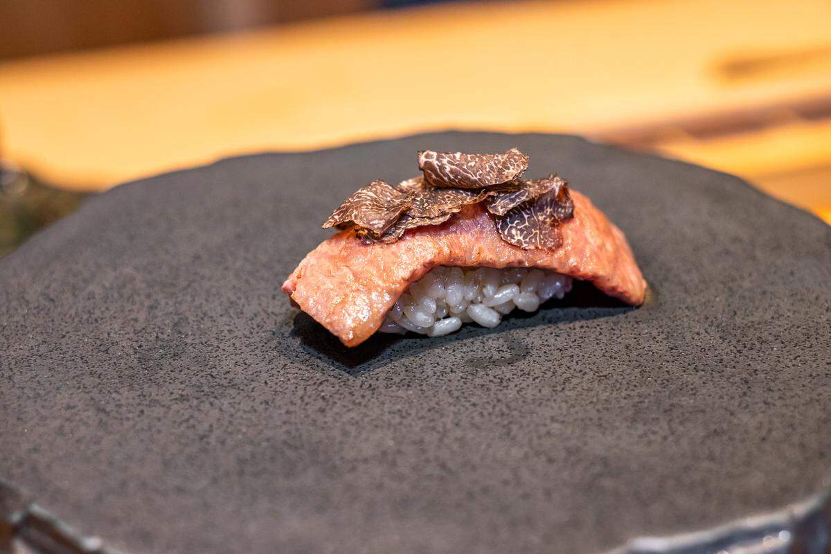 A close-up of a piece of seared beef nigiri topped with thin shavings of black truffle, served on a dark, matte, textured circular plate.