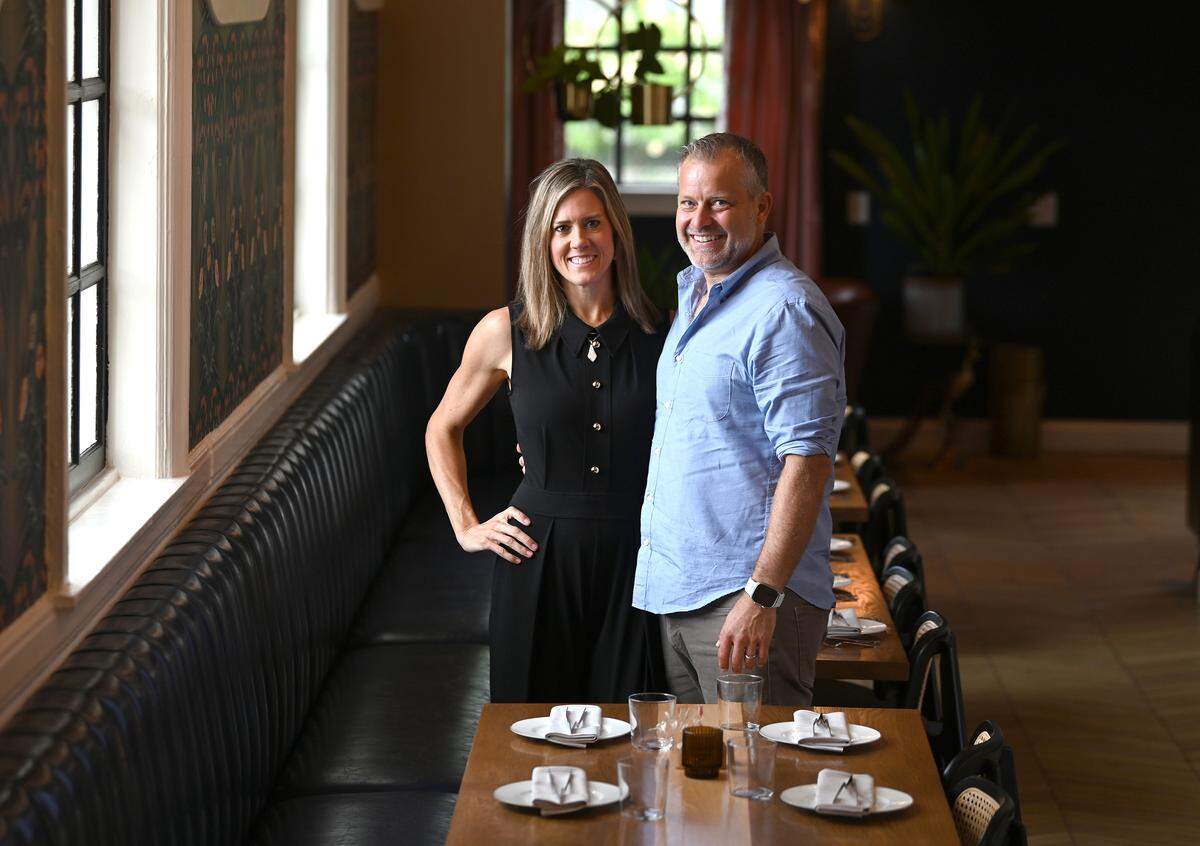 A smiling couple stand in a stylish, dimly lit restaurant dining room. The restaurateur on the left is wearing a black sleeveless dress and has a hand on the hip. The one on the right is wearing a light blue button-down shirt and grey trousers. They are positioned behind a row of set dining tables, in front of a long, black tufted leather booth that runs along a wall with tall windows and patterned wallpaper. The background features deep blue or black walls, dark red curtains, and some green plants.