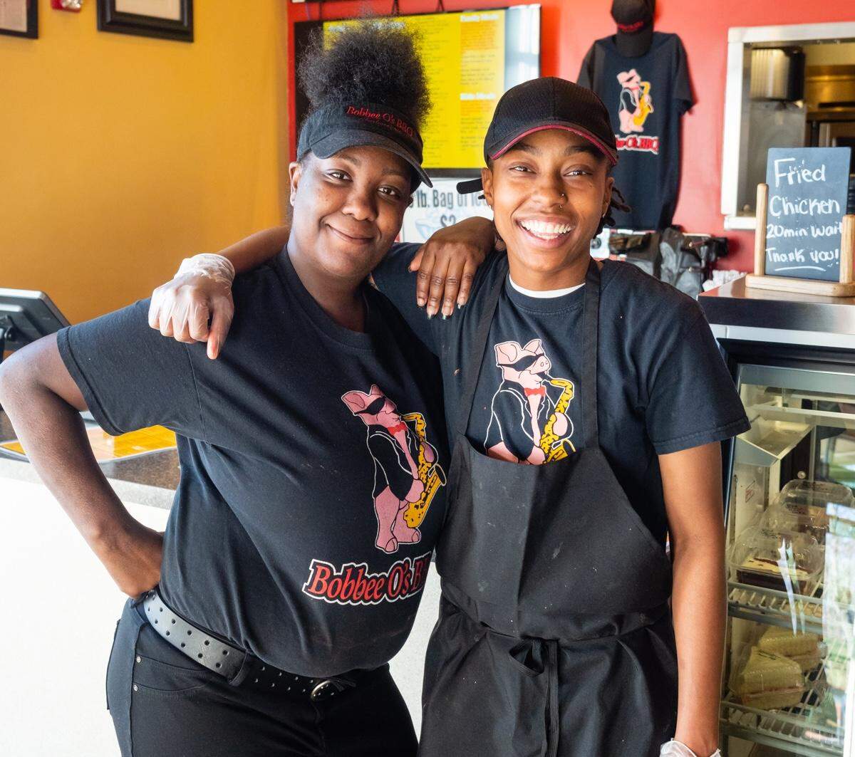 Two people standing behind a restaurant counter, smiling warmly at the camera with their arms around each other. They are wearing black t-shirts and visors featuring a cartoon pig mascot playing a saxophone.