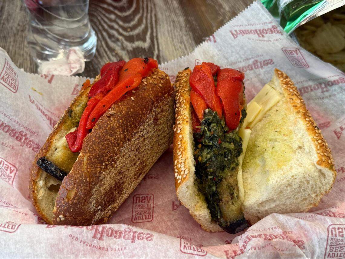 A close-up of a vegetarian hoagie sliced in half. The sandwich features grilled eggplant, bright red roasted peppers, and sautéed greens (possibly broccoli rabe) with slices of white cheese on a sesame seed roll.