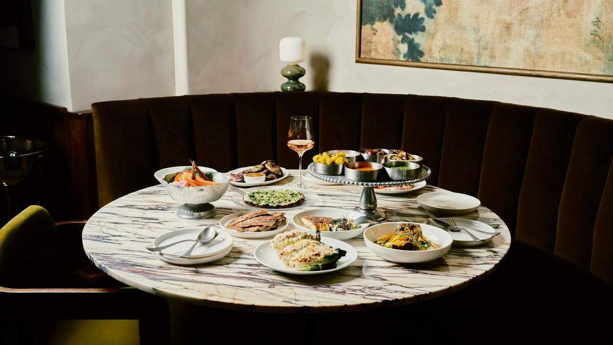 A luxurious dining table set for a meal. The round, white marble table is filled with various dishes in white and metal bowls and plates. There is a single wine glass with rosé wine. A green candle holder with a white candle sits near the back, and a framed painting with a dark blue and green floral design hangs on the wall behind the booth seating. The booth itself is upholstered in a deep brown, plush velvet.