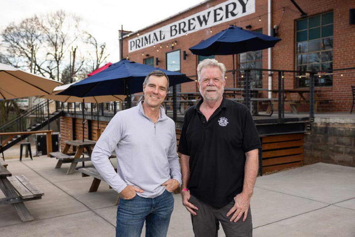 Two business owners stand smiling on the outdoor patio of Primal Brewery. The brewery’s brick exterior and patio umbrellas are visible behind them.