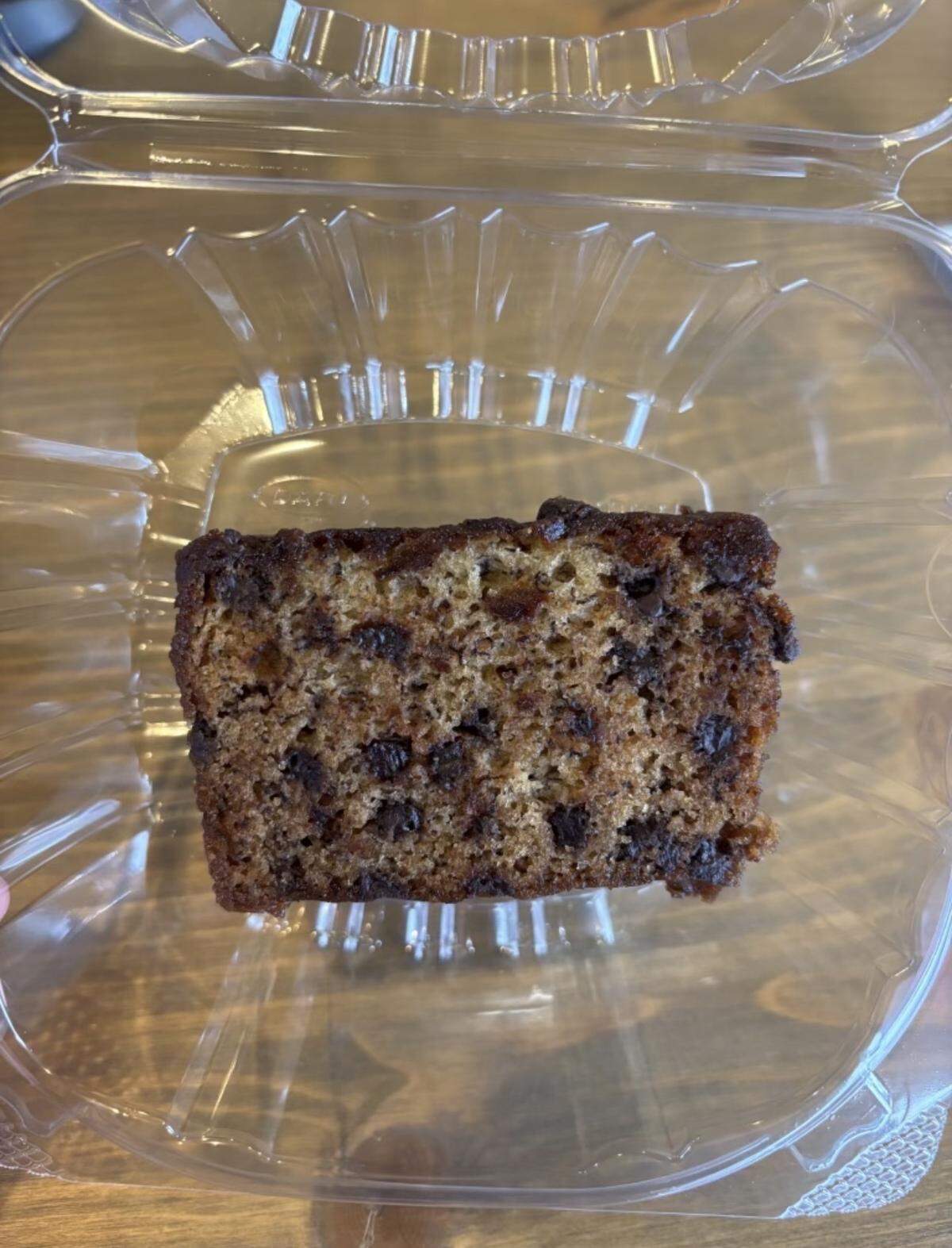 A thick slice of chocolate chip banana bread, moist and dark brown in color, sitting inside a clear, square plastic clamshell container.