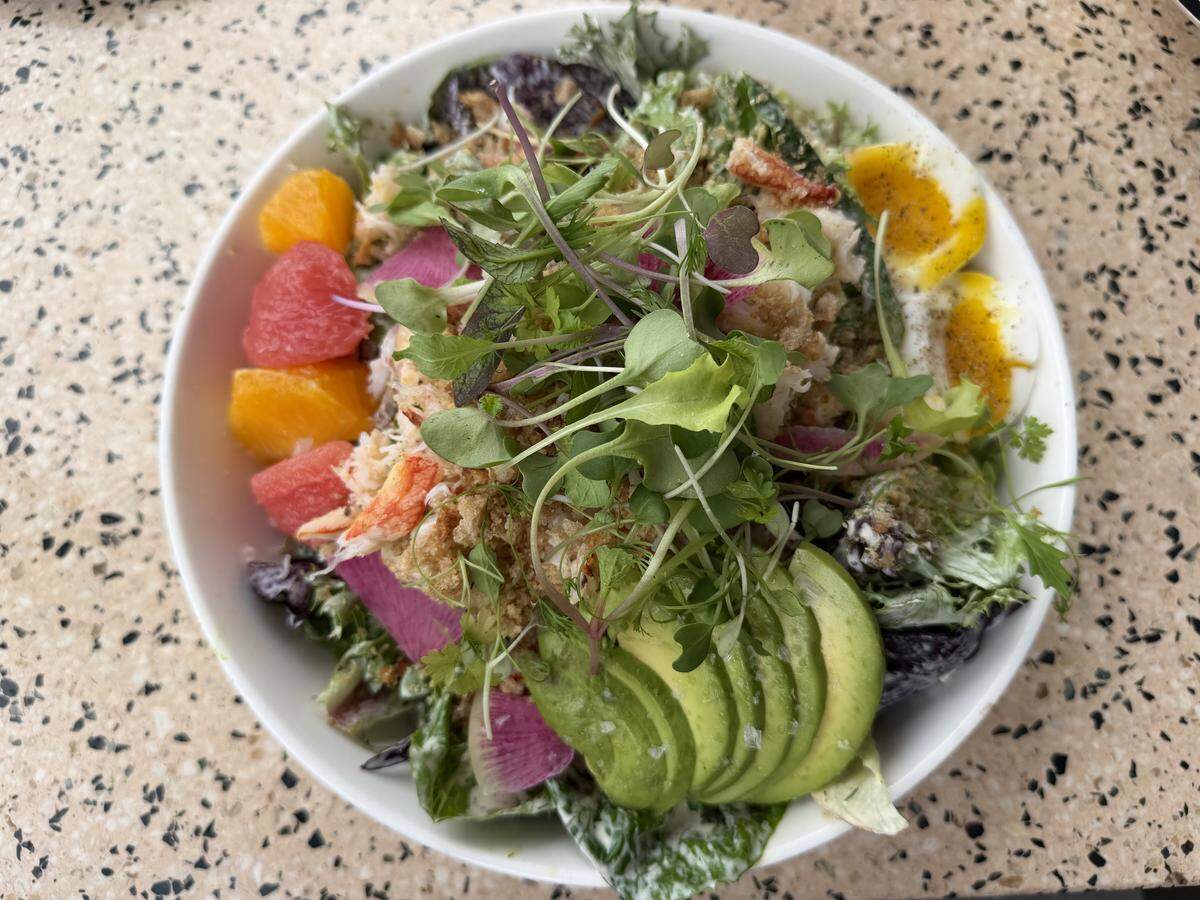 A vibrant, colorful salad bowl viewed from above. The bowl is packed with mixed greens, sliced avocado, soft-boiled egg halves topped with black pepper, watermelon radish, and segments of orange and grapefruit. Large pieces of crab meat or lobster and a handful of fresh microgreens finish the dish, which sits on a speckled granite tabletop.