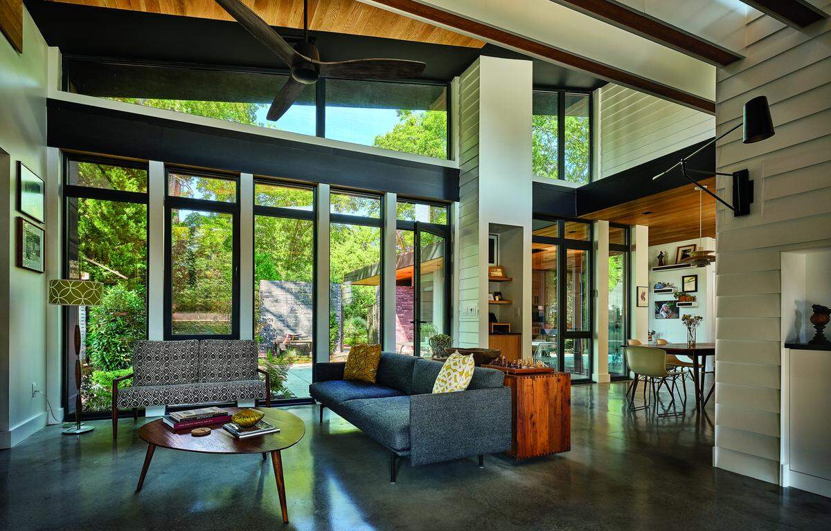 A wide shot of a modern, open-concept living room with mid-century modern aesthetics. The room features high ceilings with a large fan, polished concrete floors, and a two-story wall of black-framed windows that look out onto a lush, green yard. The space is furnished with two sofas, one gray and one patterned, and an oval-shaped wooden coffee table. In the background, a dining area is visible on a raised level.