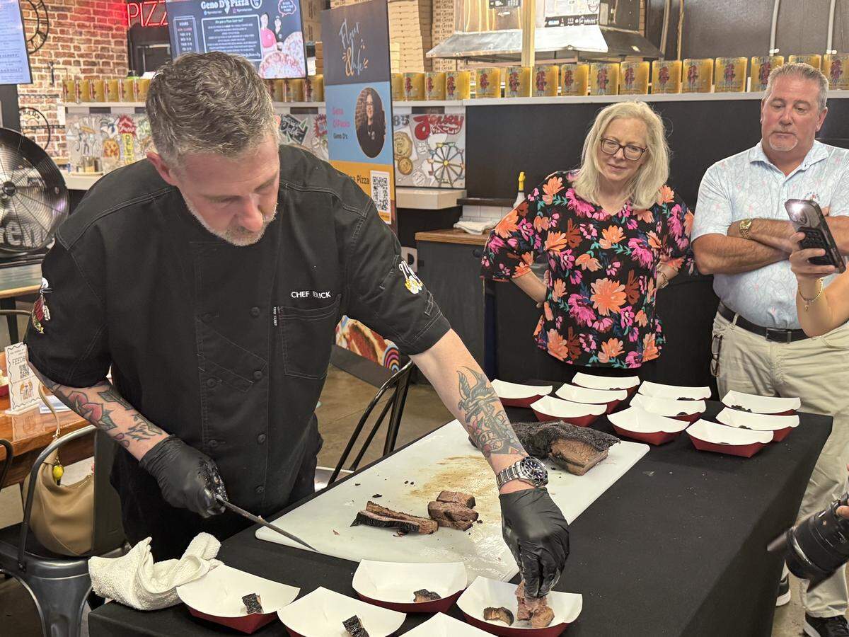 A chef wearing a black uniform embroidered with “CHEF ERICK” and black disposable gloves stands behind a black-clothed table. He has visible arm tattoos and is slicing a piece of smoked brisket on a white cutting board, placing small portions into individual white and red paper food boats. In the background, a woman in a colorful floral blouse and a man in a light blue polo shirt stand together watching the demonstration. The setting is indoors, featuring a pizza counter with a brick facade and promotional signs in the background.