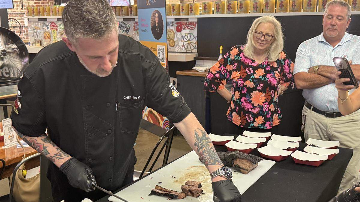 A chef wearing a black uniform embroidered with “CHEF ERICK” and black disposable gloves stands behind a black-clothed table. He has visible arm tattoos and is slicing a piece of smoked brisket on a white cutting board, placing small portions into individual white and red paper food boats. In the background, a woman in a colorful floral blouse and a man in a light blue polo shirt stand together watching the demonstration. The setting is indoors, featuring a pizza counter with a brick facade and promotional signs in the background.