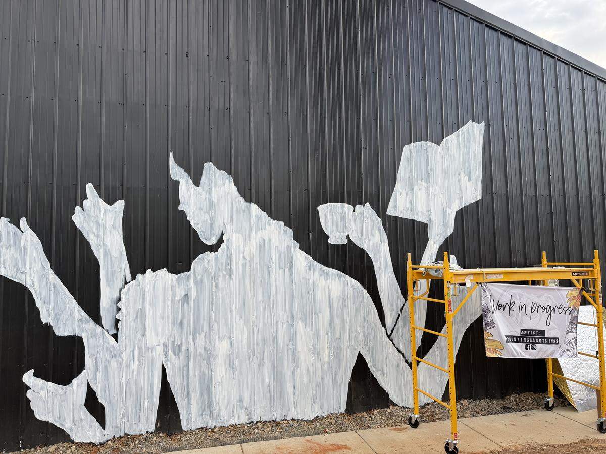 A white primer outline of a mural on a black corrugated metal wall. The mural depicts a horned, multi-armed figure holding a teacup and a book. A yellow rolling scaffold stands to the right with a “Work in Progress” banner attached, crediting the artist @paintingsandthings.