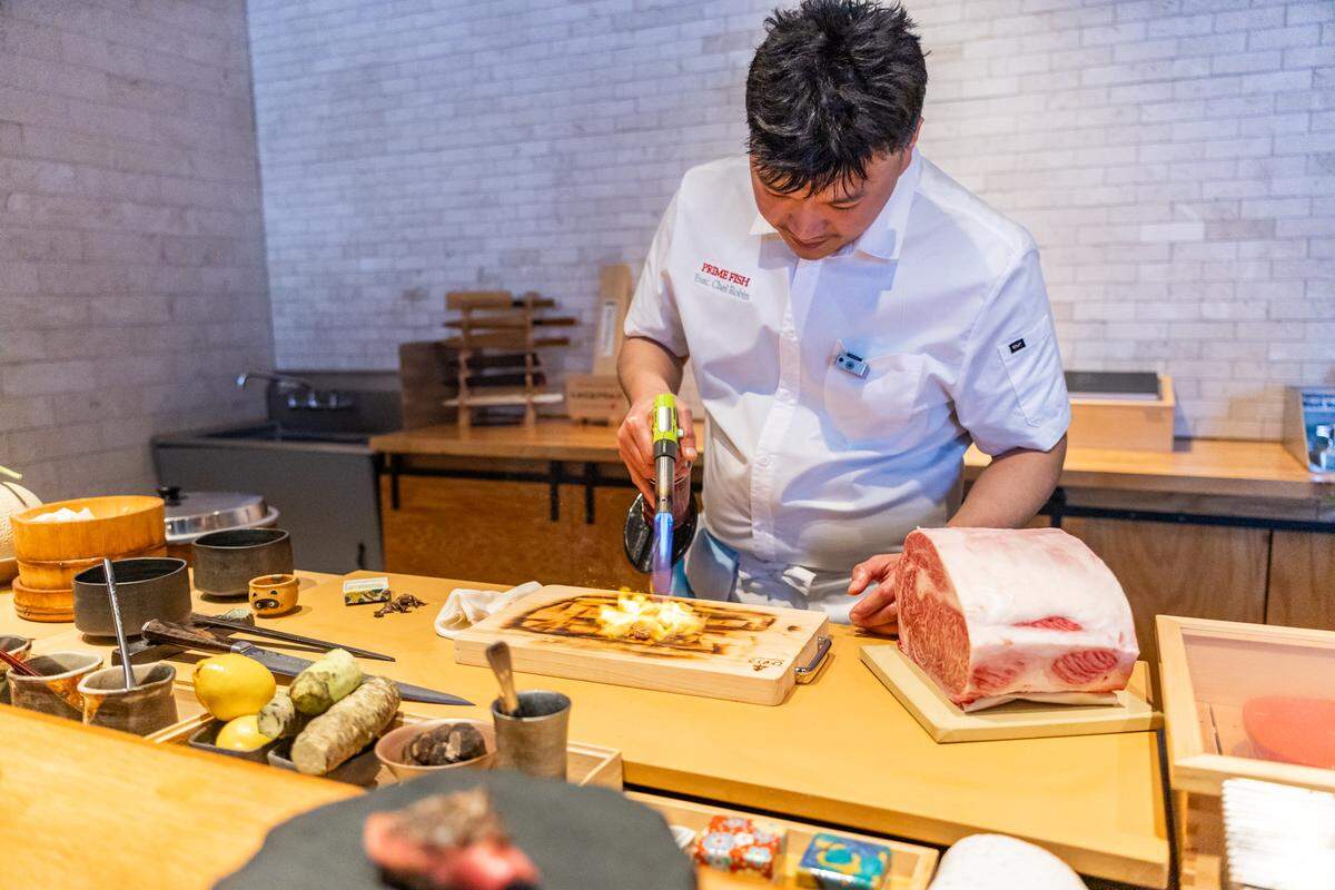 A chef uses a kitchen torch to sear a wooden board on the counter. A large, marbled slab of raw wagyu beef sits nearby on the wooden workstation, surrounded by various culinary tools and ingredients.