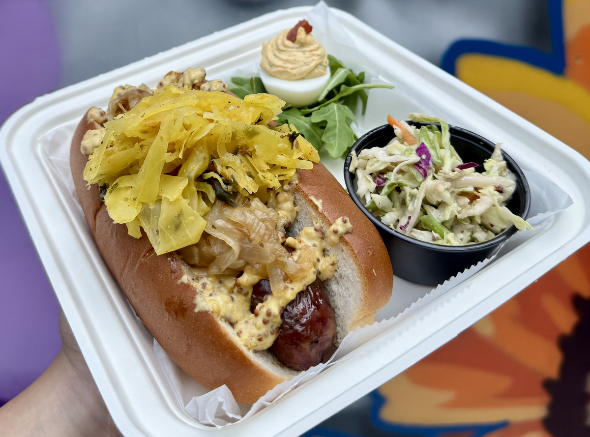 A handheld shot of a meal in a white molded-fiber container. A large beer bratwurst is in a split-top bun and is topped with mustard, onions, and sauerkraut. Accompanying the bratwurst is a small black cup of coleslaw and half a deviled egg resting on arugula. The background is a colorful blur.