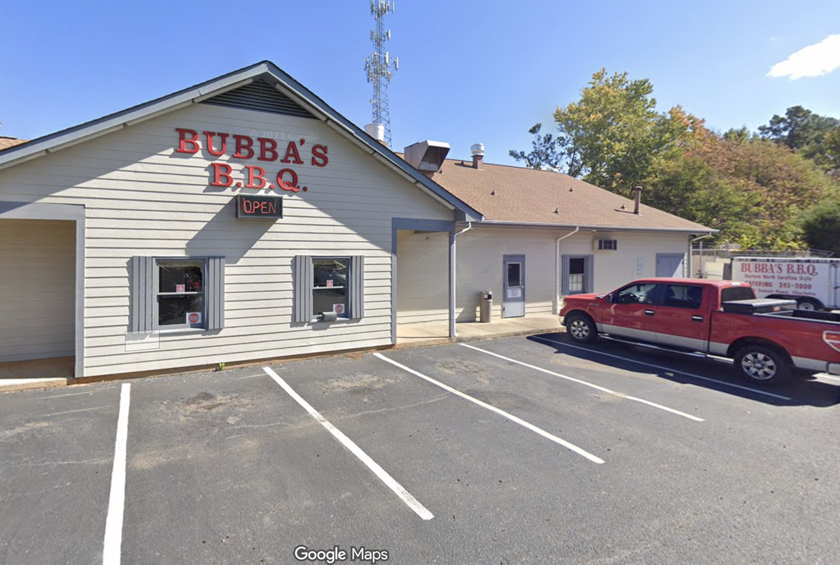 A daytime exterior shot from Google Maps shows a single-story commercial building with light gray siding with signage in red lettering on the front of the building reads “BUBBA’S B.B.Q.” with a small neon “OPEN” sign beneath it. A red pickup truck is parked in the asphalt lot in front of the building, and a white catering trailer with the business name is visible in the background. The building is flanked by green trees and a tall communication tower in the distance under a clear blue sky.