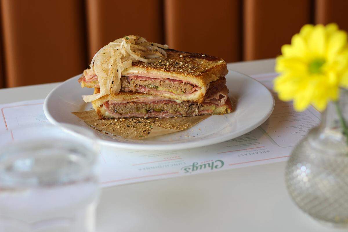 A delicious-looking, pressed Cuban-American sandwich is presented on a white plate. The sandwich is cut in half and stacked, revealing its layers of meat, melted cheese, pickles, and a thin layer of mustard. It’s topped with a pile of sautéed onions. The plate rests on a white table with a menu visible underneath. In the foreground, a glass of water and a vase with a bright yellow flower are slightly out of focus. The background is a warm, orange-brown cushioned booth.