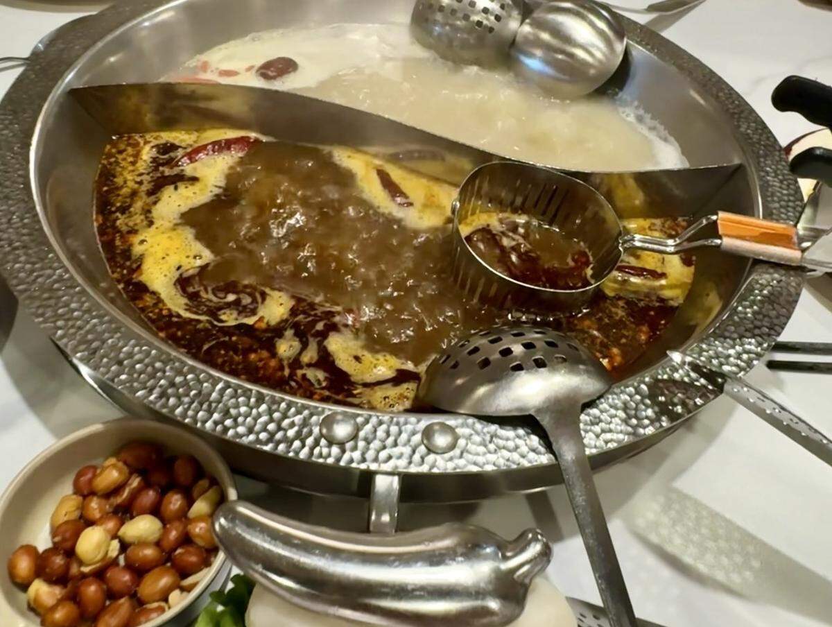 A top-down close-up of a large metal hotpot divided into two sections. One side contains a dark red, bubbling spicy broth with floating dried chilies; the other holds a creamy, pale white mild broth. Several slotted metal ladles and a small wire cooking basket are resting in the pot. Small side dishes of roasted peanuts and white buns are visible in the foreground.