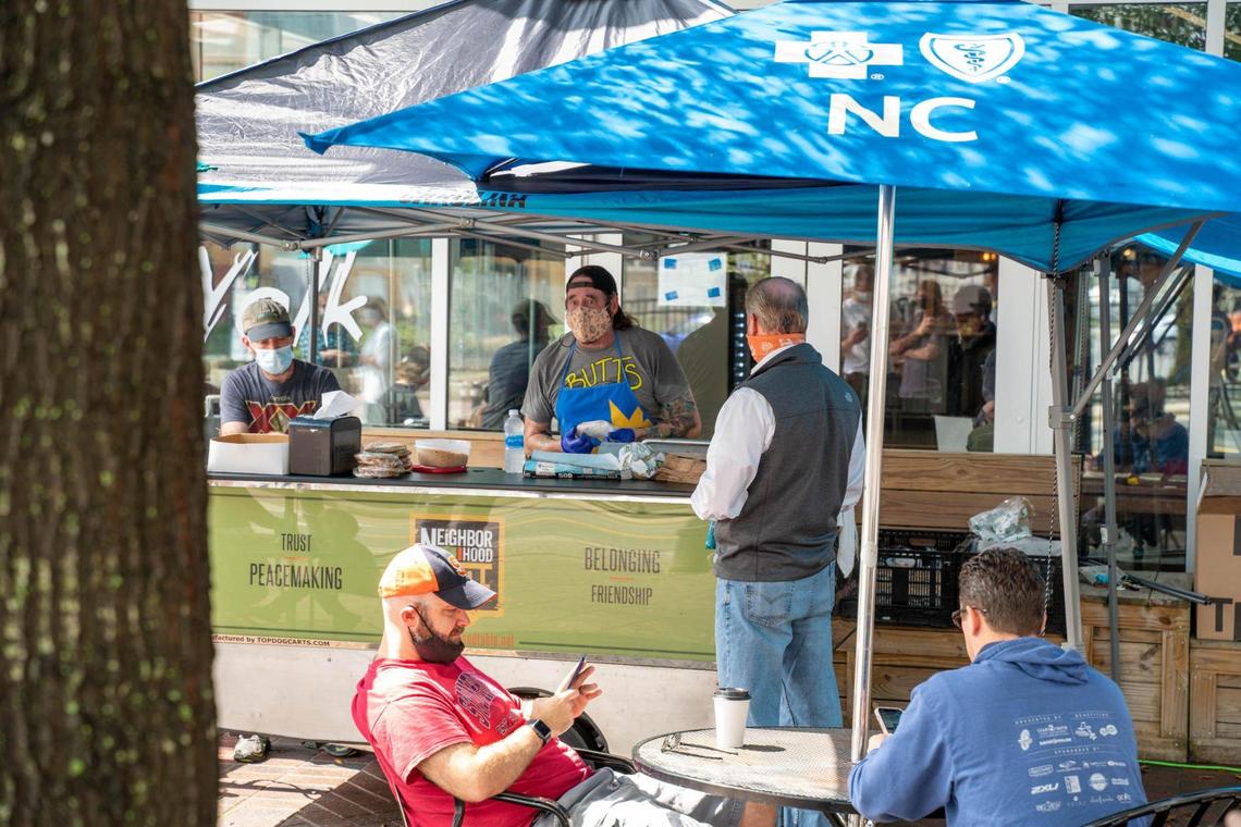 A bustling outdoor food stand scene with a person in a gray t-shirt and a blue apron serving a customer at a counter under a blue umbrella with “NC” and a cross symbol on it. The vendor is wearing a patterned face mask. In the foreground, a customer in a red t-shirt and a baseball cap is seated at a round table, looking at a phone. Another person in a blue hoodie is seated at the same table, facing away. In the background, another food stand is visible under a black awning. The scene is slightly blurred, giving a sense of movement. A sign on the counter reads “Trust, Peacemaking, Neighbor Hood, Belonging, Friendship.”