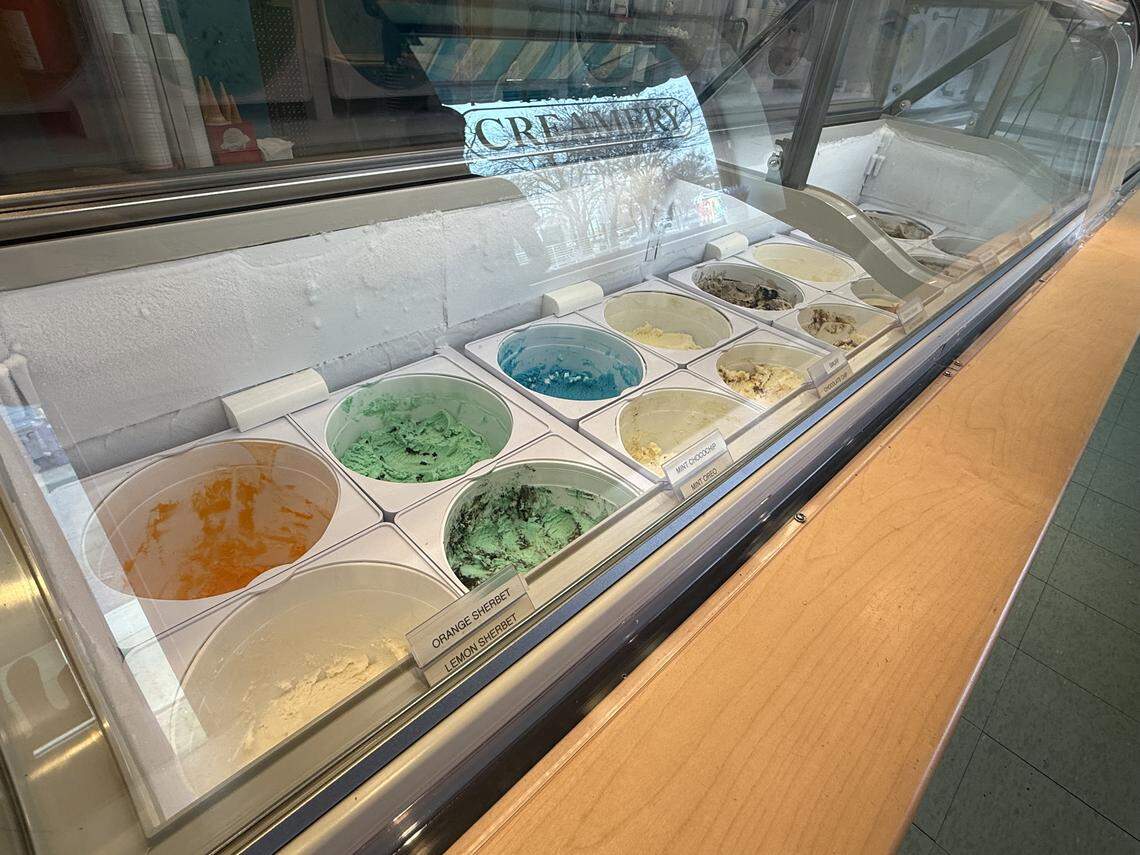 A view into a glass display freezer showing several tubs of ice cream and sherbet, including bright orange, mint chocolate chip, and lemon flavors.