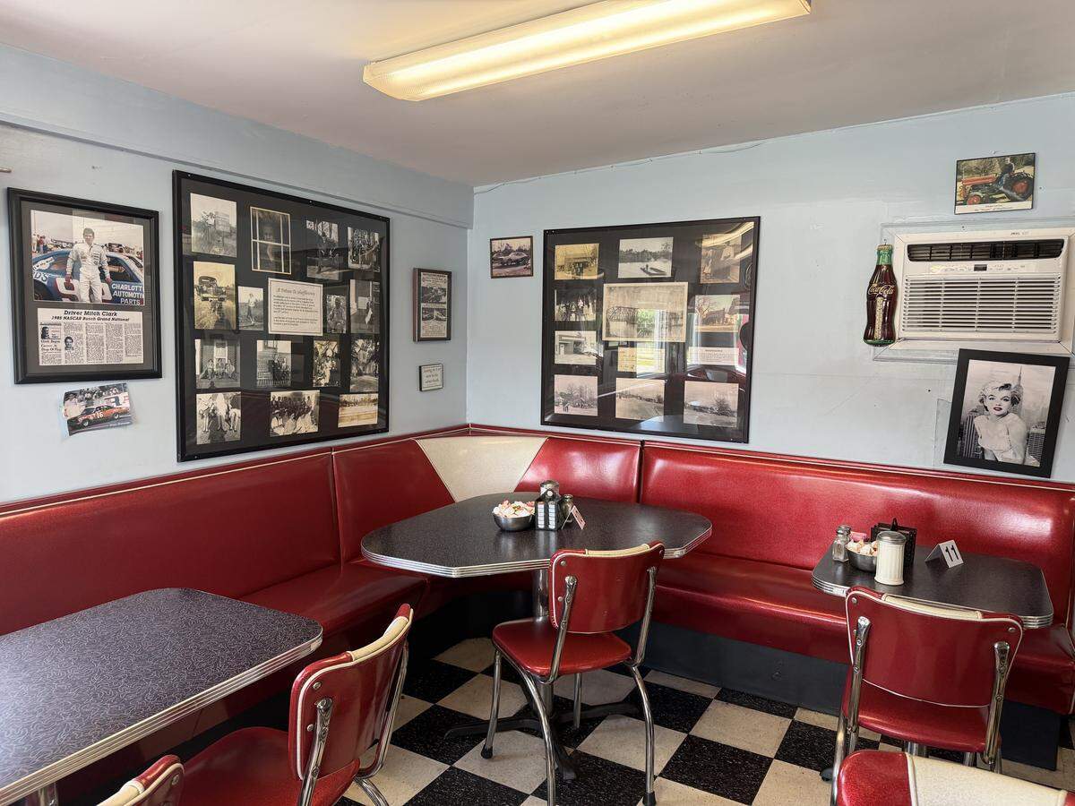 A nostalgic corner booth in a diner featuring bright red vinyl seating with white “V” accents. Two grey speckled tables are paired with red-and-chrome chairs over a black-and-white checkered floor. The light blue walls are covered in framed memorabilia, including local historical photographs, a NASCAR tribute, and a portrait of Marilyn Monroe. A vintage-style Coca-Cola bottle is mounted near a small window air conditioning unit.