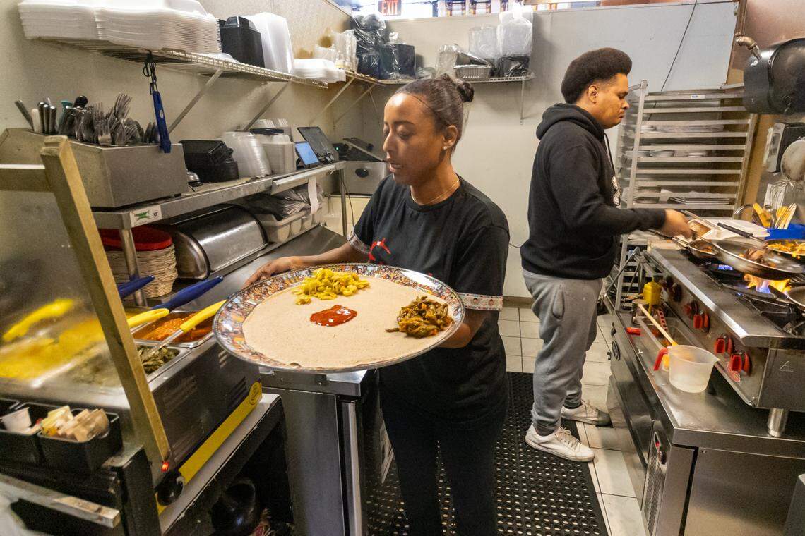 A busy kitchen scene where a restaurateur in a black “Enat” t-shirt is carefully preparing a large communal platter while holding a silver tray topped with a wide circle of Injera (traditional sourdough flatbread) and adding portions of colorful stews, including yellow split peas and greens. In the background, a person in a black hoodie works at a commercial stove with flames visible under sauté pans, while shelves are stocked with takeout containers.