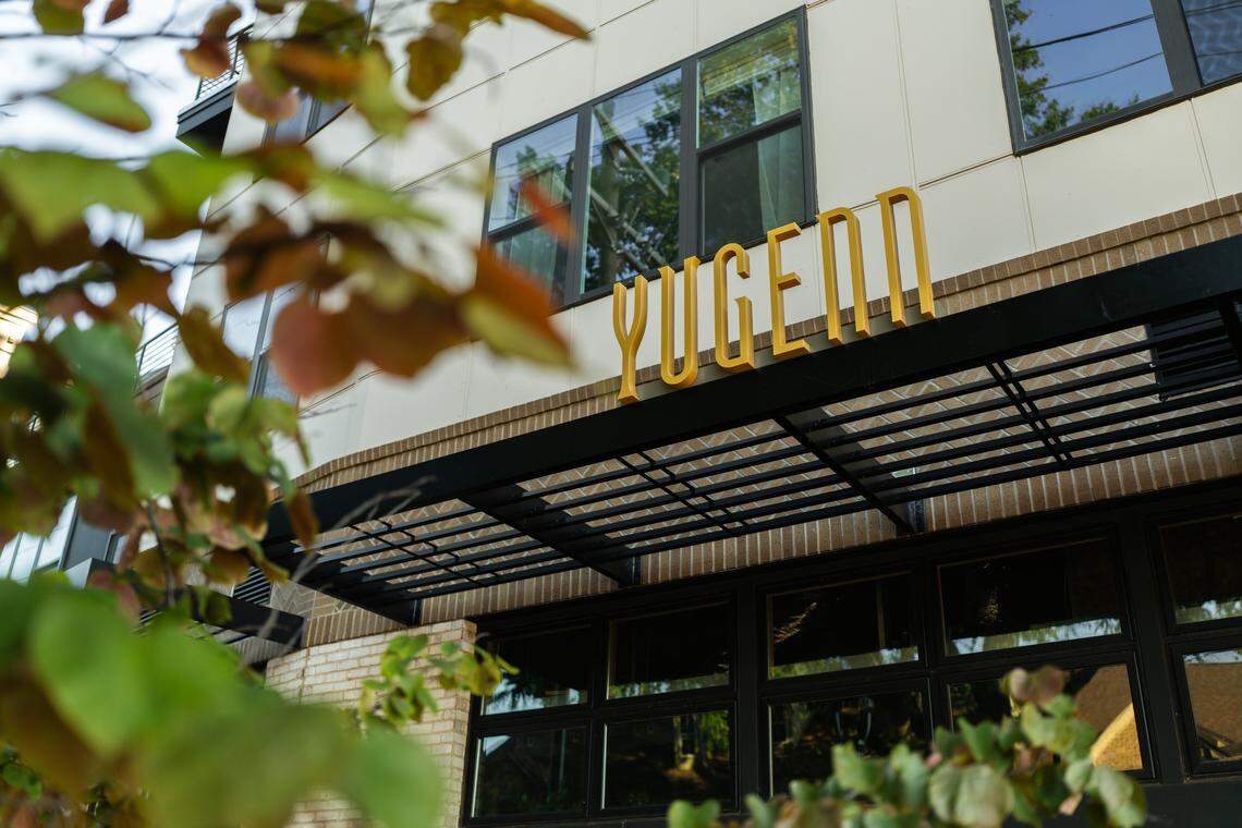 A low-angle shot of the front of a modern building with a large yellow sign that reads YUGENN mounted on a brick wall. The building has large windows, and a black metal awning with a grate-like pattern extends over the entrance below the sign.