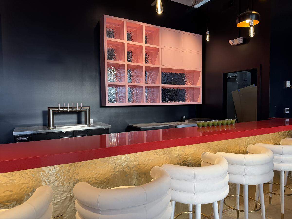 A modern bar interior featuring a bright red countertop and a gold hammered-metal base. White, tiered swivel barstools line the counter. On the dark matte wall behind the bar, a large pink grid shelving unit with textured glass inserts serves as a focal point next to a stainless steel draft beer tower.