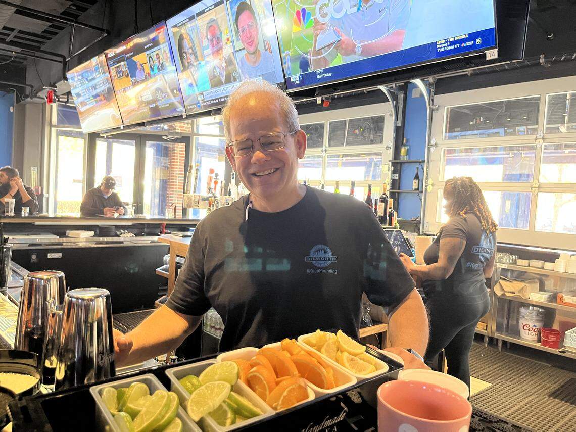 An eye-level, medium shot of a smiling man with short gray hair and glasses, wearing a black t-shirt, as he stands behind a bar holding a white tray filled with lime, orange, and lemon slices. In the background of the bright, open-air bar, multiple televisions are mounted above, and large, open garage-style doors are visible.