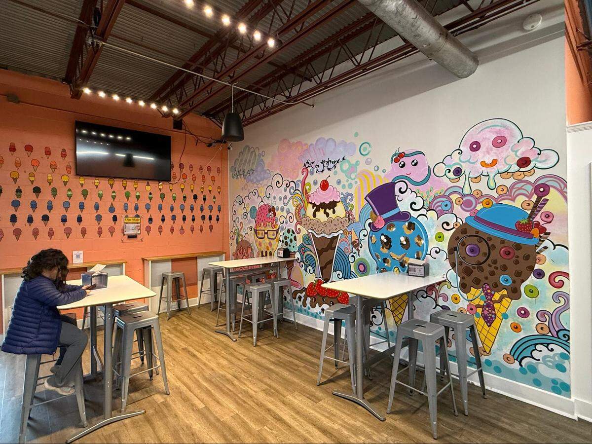 An ice cream shop’s seating area featuring high-top white tables and silver metal stools. One wall is covered in a vibrant, psychedelic mural of cartoonish ice cream characters, while another orange wall is decorated with dozens of small, colorful ice cream cone illustrations.