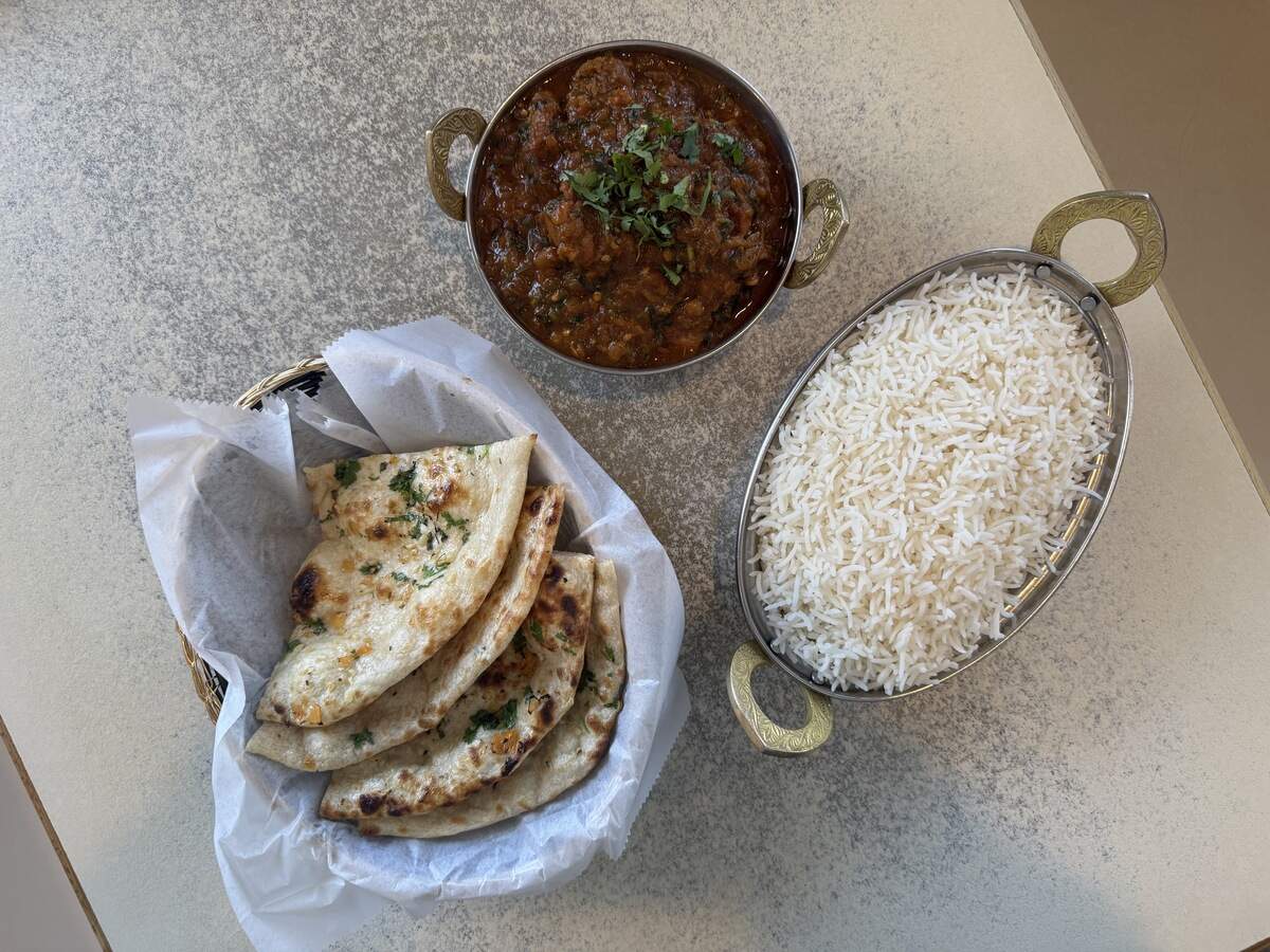 A top-down view of an Indian meal arranged on a speckled light-colored tabletop. At the top sits a round, metal serving dish filled with a rich, reddish-brown curry garnished with fresh chopped cilantro. To the right is an oval metal dish piled high with white basmati rice. On the left, a woven basket lined with white paper holds several pieces of golden-brown garlic naan sprinkled with herbs.