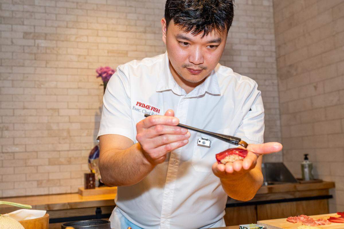 A chef uses a small brush to apply a glaze to a. handheld piece of wagyu, focusing on the work against a background of a light-colored brick wall.