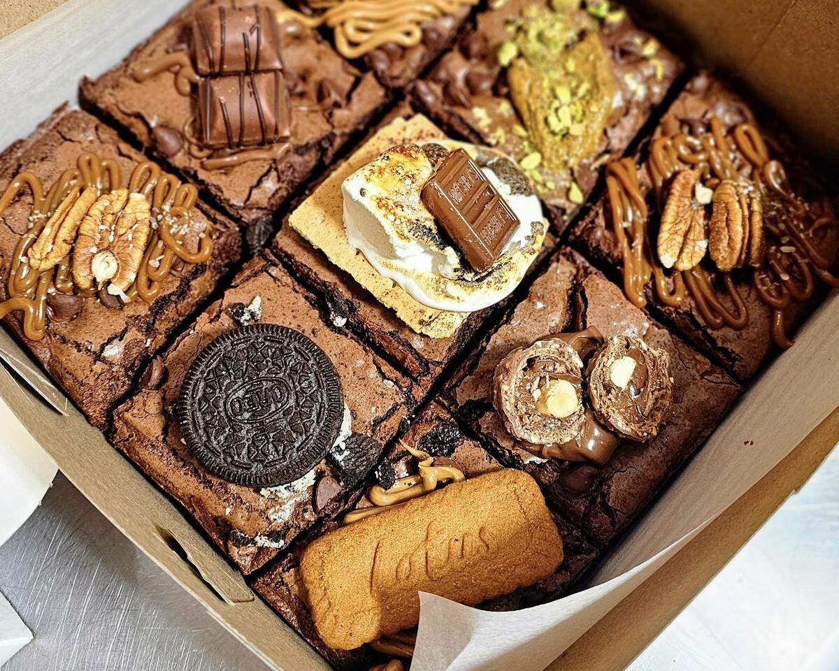 A 3x3 grid of nine decadent, fudgy brownies is displayed in a cardboard box. Each brownie is a different flavor, showcased by its topping: one is a s’mores brownie, another is topped with an Oreo, one with a Biscoff cookie, one with pecans and caramel, one with pistachios, and others with various chocolate candies.