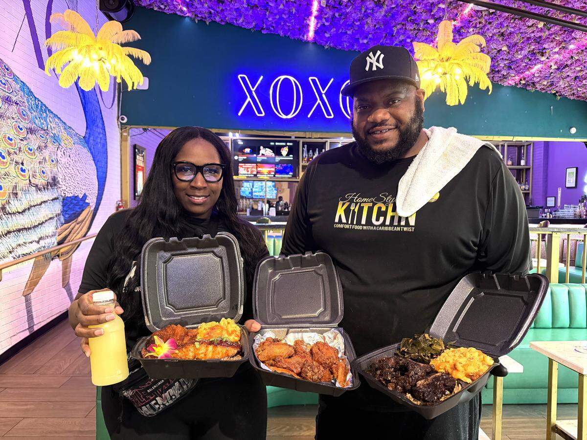 A medium shot of a smiling couple holding open takeout containers of comfort food inside a brightly decorated restaurant. The person on the right wears a black baseball cap and a logo t-shirt, while the one on the left wears glasses and holds a bottled beverage. Their containers feature glazed salmon, fried wings, oxtail, macaroni and cheese, and greens. The vibrant background includes a neon “XOXO” sign, teal walls, and purple floral ceiling decorations.